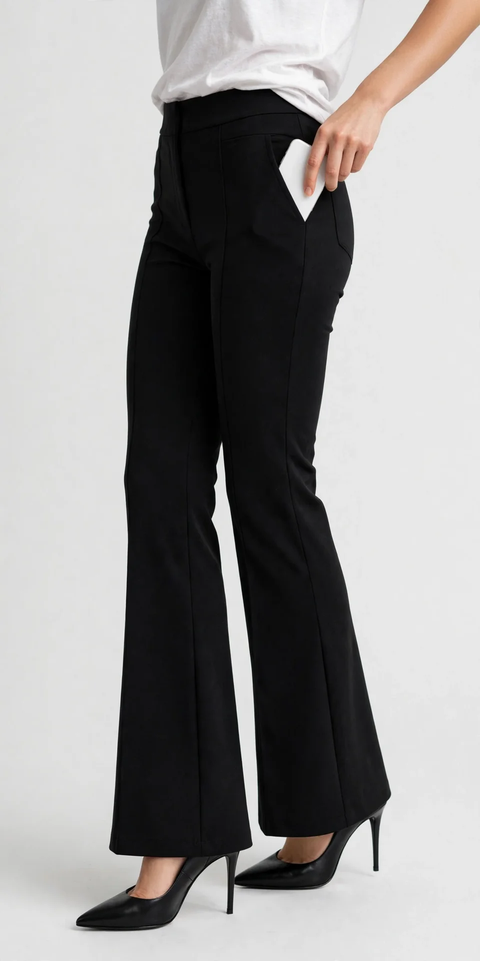 Cropped torso-down model wearing a white top and black high-waist flare pants with a front seam, holding a white phone in a side pocket, wearing black pointed stiletto heels