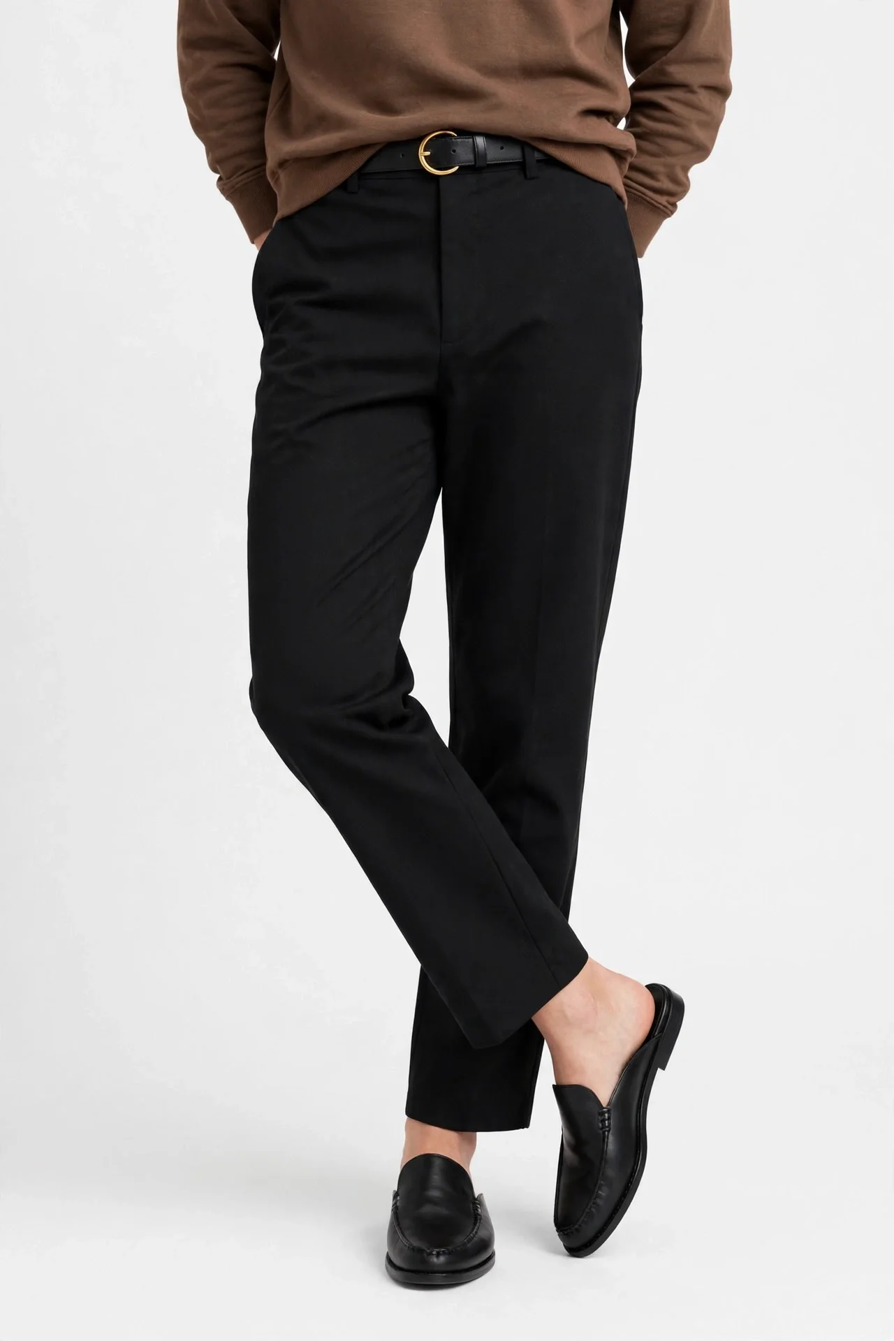 Black straight-leg pants worn with a brown long-sleeve top, thin black belt with gold round buckle, and black mule-style loafers, one leg crossed in front of the other