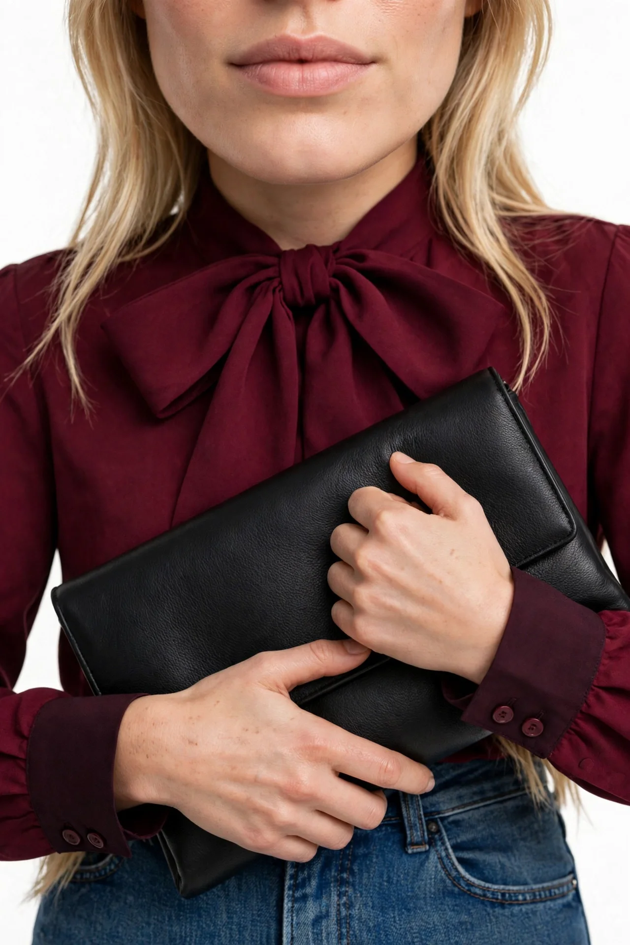 Blonde model wearing a deep burgundy bow-tie blouse with a large bow at the neckline and button cuffs, with blue jeans, holding a black clutch