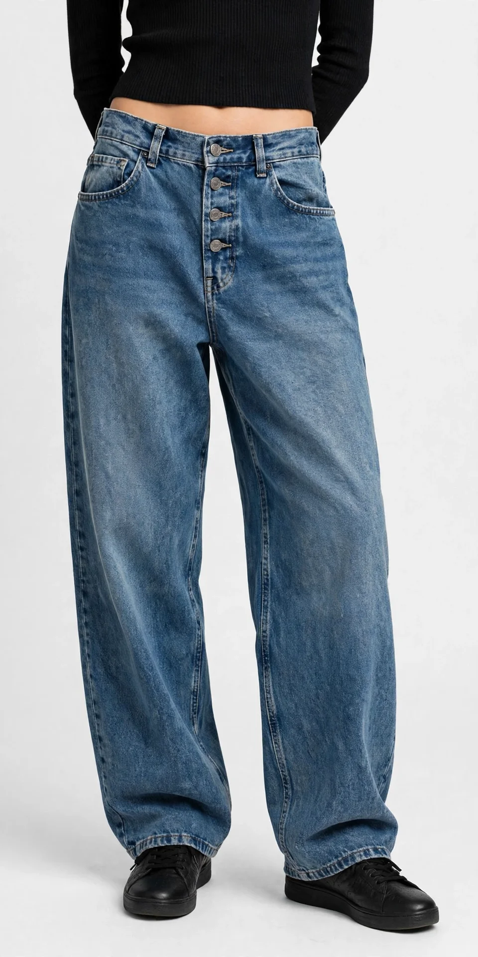 Model shown waist-down wearing a cropped black top, medium-wash blue baggy wide-leg jeans with a high waist and button fly, and black shoes