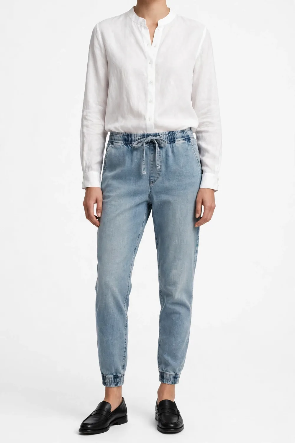 Model from the neck down wearing a white blouse, light faded-blue tapered jogger pants with elastic cuffed ankles, and black loafers, on a white background