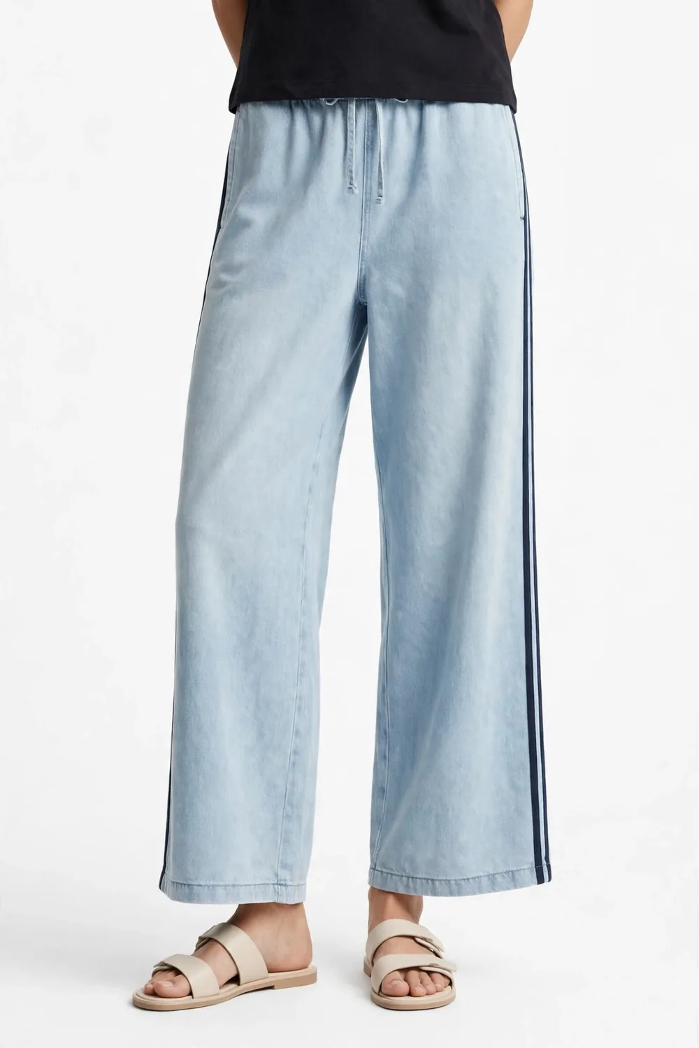 Model from the waist down wearing a dark top, light-wash wide-leg track pants with a dark stripe down the outer side of each leg, and light-coloured flat sandals, on a white background