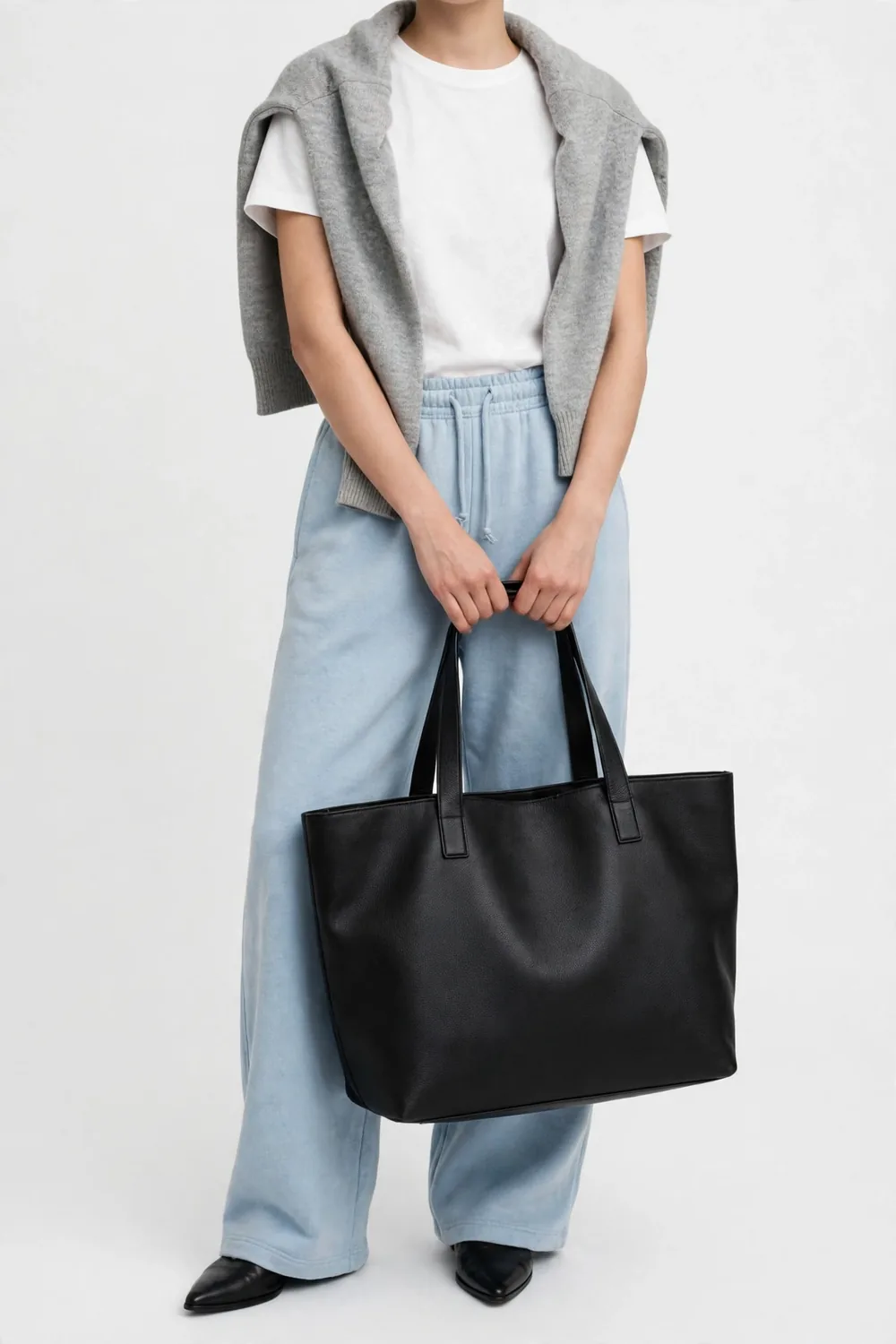 Model in a white t-shirt with a grey sweater draped over the shoulders, wearing very light-blue wide-leg jeans-like pants and black pointed shoes, holding a large black bag, on a white background