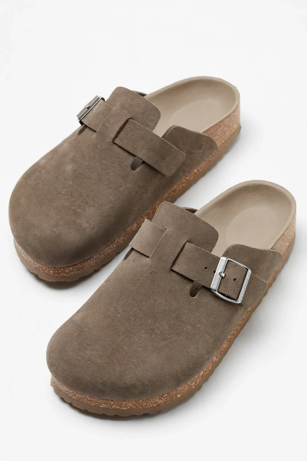 Pair of taupe suede clogs with wide buckle straps and a cork-style footbed on a white background