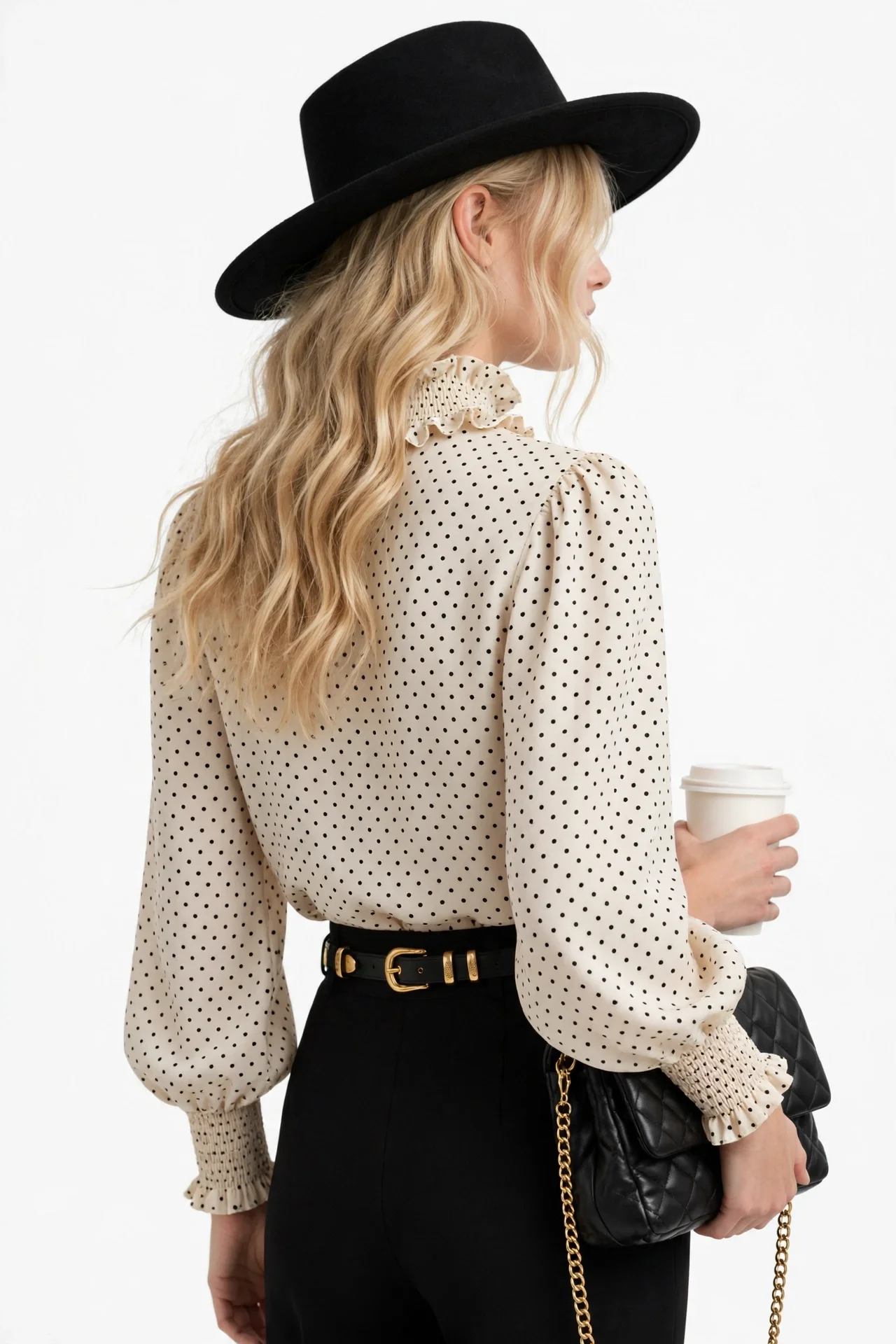 Blonde model in a black wide-brim hat wearing a cream polka-dot blouse with ruffled mock neckline and smocked cuffs, black trousers, holding a coffee cup and a black chain clutch
