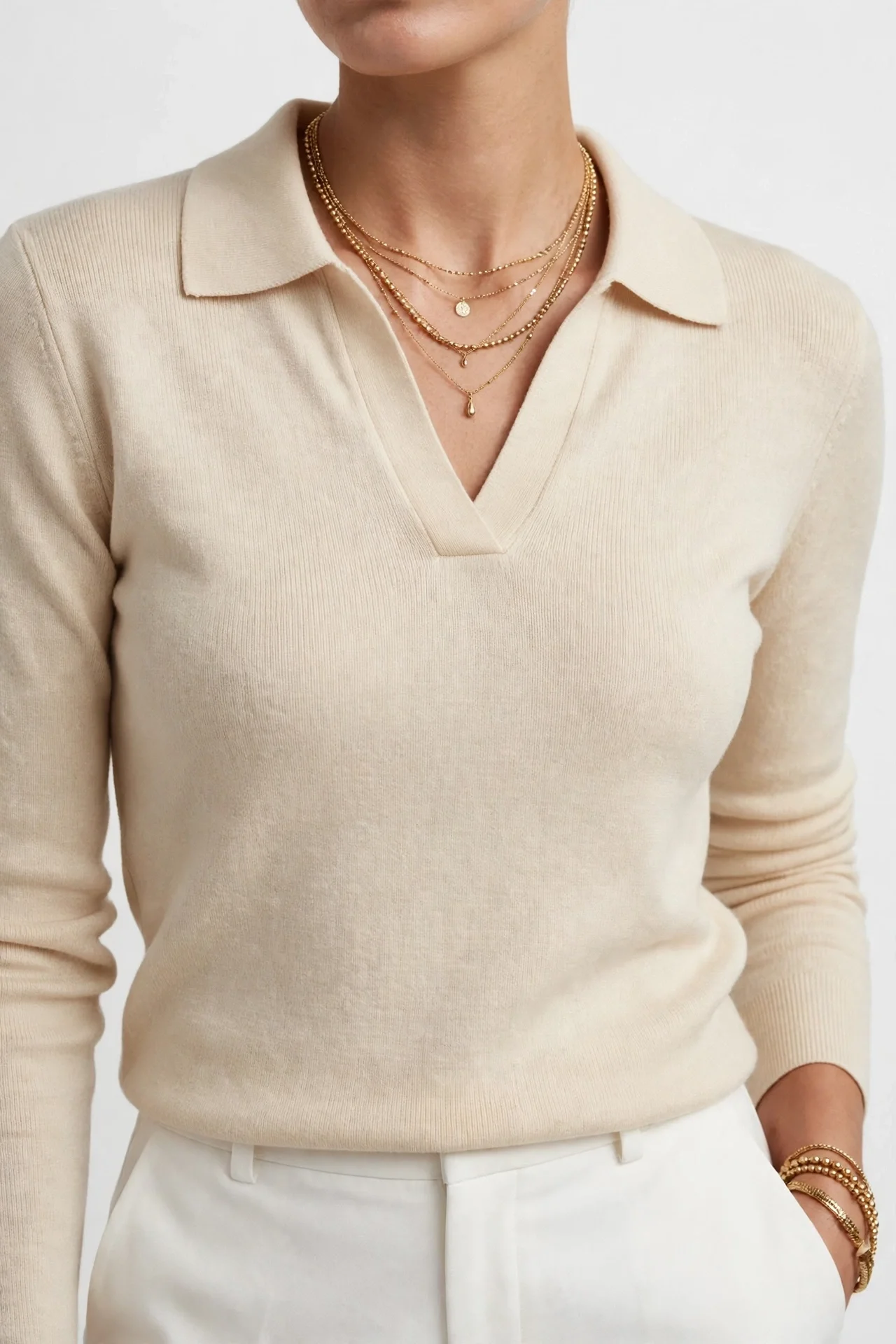 Beige cream polo sweater with fold-over collar and V-neck, worn with white trousers and layered necklaces