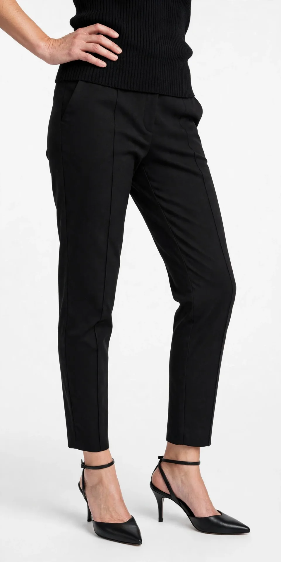 Black tapered ankle-length pants with a visible centre pintuck seam down the front of each leg, worn with a black ribbed top, one hand on the hip, and black pointed-toe heeled pumps with ankle straps