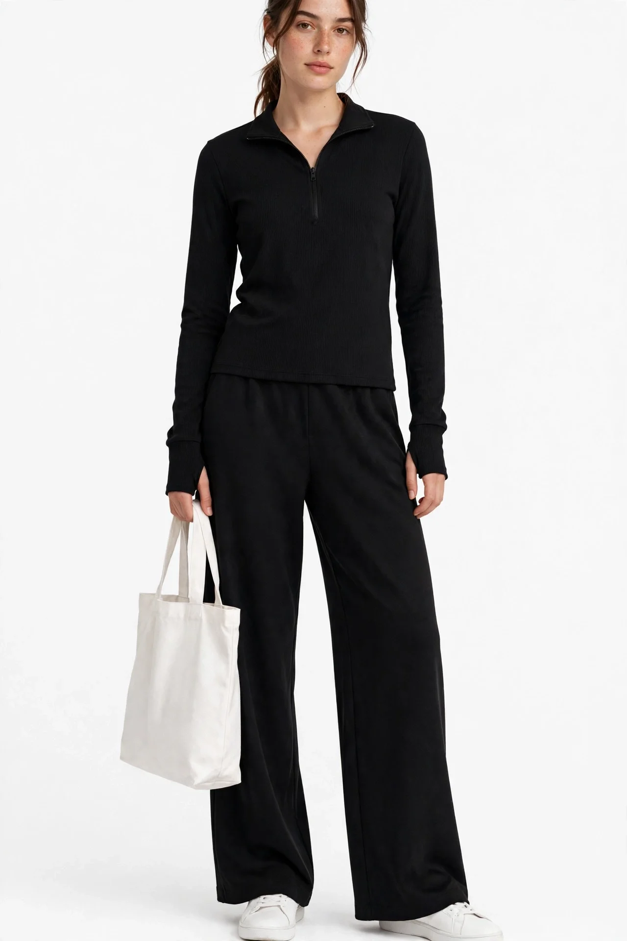 Model wearing a black half-zip long-sleeve top and matching black wide-leg pants, white shoes, holding a white bag