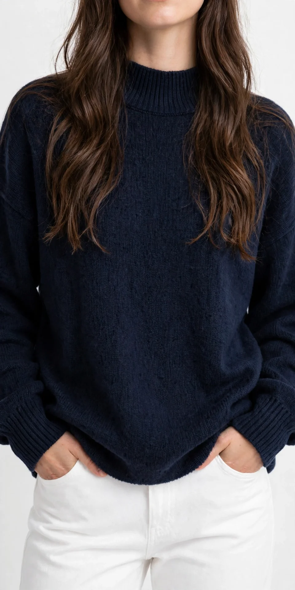 Woman's torso with long brown hair, wearing a dark navy oversized mock-neck sweater with ribbed collar and cuffs, hands in white pants pockets