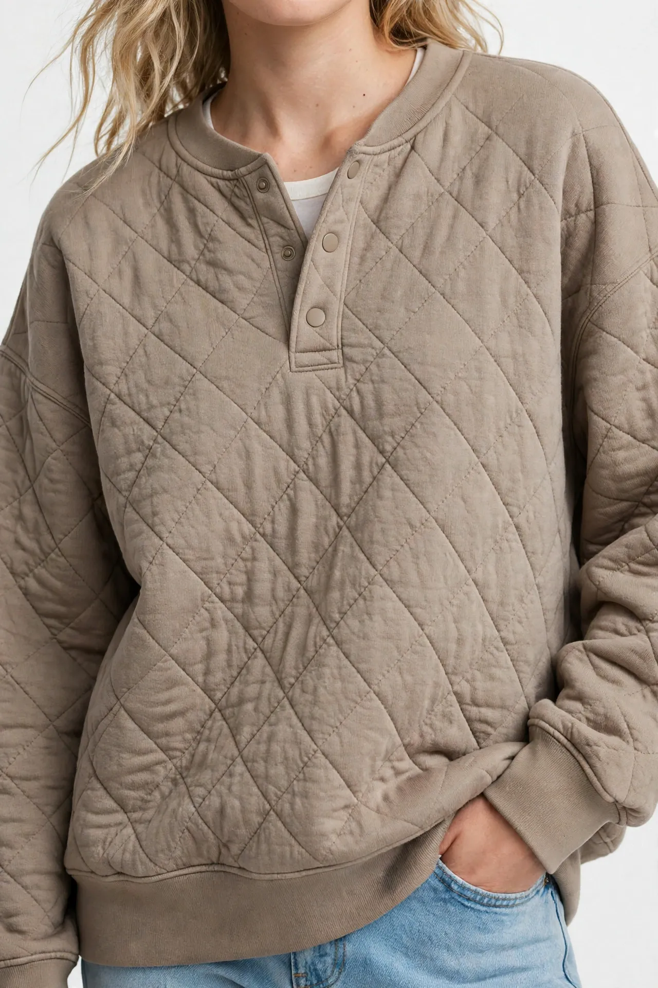 Torso view of a model with partly visible blonde hair, wearing an oversized beige taupe sweatshirt with a snap-button Henley placket and quilted diamond detailing, a white undershirt, and light blue jeans, with one hand in a pocket